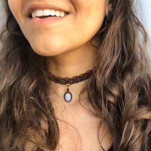 K&ouml;nnte beinhalten: Eine braune Makramee-Halskette mit Anh&auml;nger. Die Halskette besteht aus gewebtem braunem Kordel und hat einen kleinen, oval geformten Anh&auml;nger mit einem hellblauen Stein. Die Halskette wird um den Hals getragen.