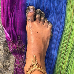 Puede incluir: Un pie cubierto de arena, con una tobillera dorada. El pie descansa sobre una toalla de playa a rayas con colores morado, azul y verde. La piel tiene un tatuaje que dice "los oceanos soy".