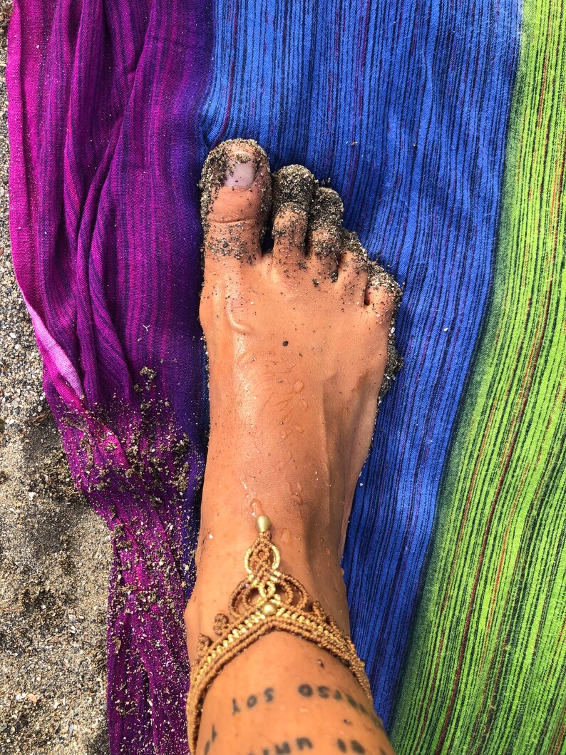 Puede incluir: Un pie cubierto de arena, con una tobillera dorada. El pie descansa sobre una toalla de playa a rayas con colores morado, azul y verde. La piel tiene un tatuaje que dice "los oceanos soy".