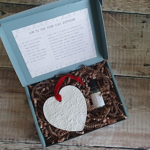 May include: A white ceramic heart-shaped diffuser with a floral design, a small bottle of essential oil labelled "Unwind", and instructions on how to use the diffuser in a blue box with brown shredded paper.