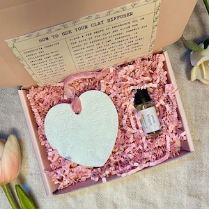 Op de afbeelding: Een hartvormige witte klei diffuser met een bloemenpatroon, verpakt in een roze doos met roze papier. Inclusief een flesje pioenrozen boeket geurolie. Instructies staan op het deksel.