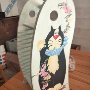 May include: A wooden magazine rack with a light green finish and a hand-painted black and white cat wearing a blue plaid scarf. The cat is surrounded by pink and yellow floral accents. The rack features a circular cutout handle at the top.