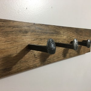 May include: A wooden coat rack with three large metal nail head hooks. The wood has a warm brown tone with visible grain. The metal hooks are a dark grey colour. The rack is designed for hanging coats.