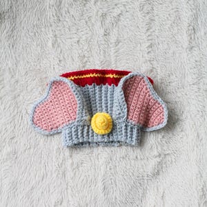 A beginner-friendly crochet pattern for a Flying Elephant dog snood. The elephant dog snood is grey with pink ears. It's wearing a tiny yellow hat and a red and yellow collar.