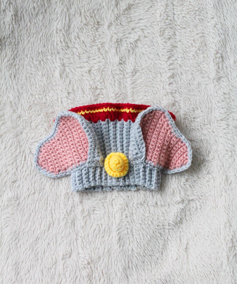 A beginner-friendly crochet pattern for a Flying Elephant dog snood. The elephant dog snood is grey with pink ears. It's wearing a tiny yellow hat and a red and yellow collar.