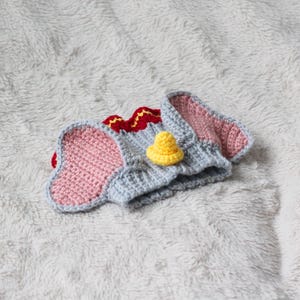 A beginner-friendly crochet pattern for a Flying Elephant dog snood. The elephant dog snood is grey with pink ears. It's wearing a tiny yellow hat and a red and yellow collar.