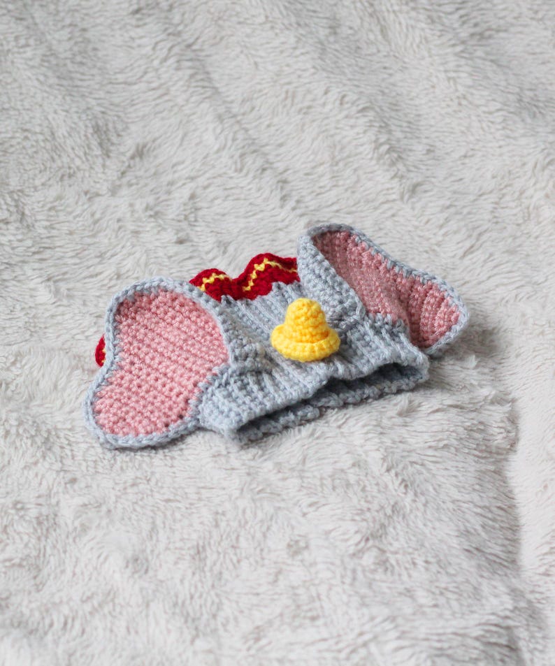 A beginner-friendly crochet pattern for a Flying Elephant dog snood. The elephant dog snood is grey with pink ears. It's wearing a tiny yellow hat and a red and yellow collar.