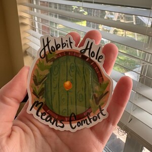 以下が含まれることがあります： 「Hobbit Hole Means Comfort」の文字が黒い筆記体で書かれた、緑色のホビットのドアが描かれたステッカー。 ドアは緑の葉のアクセントが付いた茶色のアーチに収められています。 ステッカーは手で持たれています。