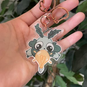 May include: A green and blue cartoon character with large eyes holding a light bulb. The character is attached to a rose gold key chain.