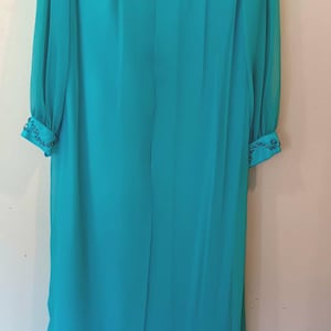 May include: A long, flowing turquoise dress with long sleeves. The neckline and cuffs are embellished with intricate beadwork. The dress is made of a lightweight, sheer fabric, and hangs on a hanger against a neutral wall.