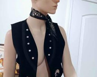 May include: A black suede vest with silver-colored button details and decorative metal accents. The vest is displayed on a mannequin, paired with a black and white paisley scarf and a white cowboy hat.