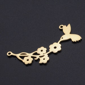 Op de afbeelding: Een goudkleurige metalen bedel met een kolibrie en bloemen. De kolibrie wordt in vlucht afgebeeld, reikend naar de bloemen op een tak. De bedel heeft een lus om aan sieraden of andere knutselwerken te bevestigen.