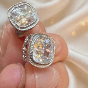 May include: A pair of silver-toned stud earrings featuring large, clear, cushion-cut stones. Each stone is surrounded by a textured, milgrain-style metal border, with an openwork gallery design visible on the sides of the setting.
