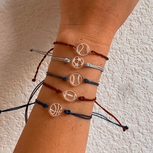May include: Five bracelets with different sports ball charms. The charms are clear with white outlines of a baseball, basketball, football, soccer ball, and tennis ball. The bracelets are made of dark blue, light blue, and brown cord.