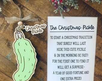 May include: A white card with black text that reads "The Christmas Pickle" and explains the tradition of hiding a pickle ornament on the Christmas tree. The first person to find it gets a surprise, a year of good fortune, and an extra prize. A green pickle ornament with a brown stem is shown on a wooden surface.