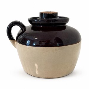 May include: A two-toned stoneware crock featuring a dark brown glazed upper section and a light beige glazed lower section. The vessel includes a single side handle, a matching dark brown lid, and a small cork stopper centered on the lid top.