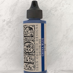 May include: A blue glass bottle of "China Blue" decor ink by Iron Orchid Designs. The bottle has a black cap and a label with ornate black lettering and a blue stripe. The bottle contains 2 fl oz of ink.