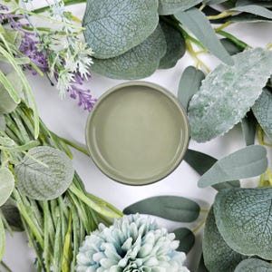 May include: A round, light green container surrounded by various green and purple foliage. The container is centered and filled with a matching light green substance. The foliage includes eucalyptus leaves, lavender, and a blue-green flower.