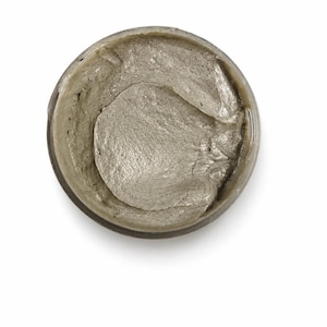 May include: A circular container filled with a thick, metallic silver paste. The substance has a dense, creamy consistency with visible swirls and ridges on the surface, reflecting light across its textured, shimmering, and opaque greyish-silver finish.