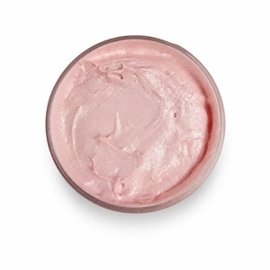 May include: A circular container filled with a thick, pale pink cream. The surface of the cream has a smooth, swirled texture with subtle peaks and valleys, contained within a clear, round vessel viewed from directly above against a white background.