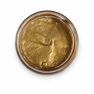 May include: A circular glass jar filled with a thick, metallic gold paste. The substance has a smooth, reflective surface with visible swirls and indentations from application, contained within a clear, transparent glass container viewed from above.