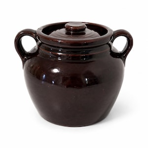 May include: A dark brown glazed ceramic bean pot with a rounded body, two side handles, and a matching lid with a central knob. The vessel features a smooth, reflective finish and a wide opening, showcasing a classic, functional kitchen design.