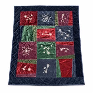 May include: A rectangular quilted throw blanket featuring a patchwork design of velvet squares in deep red, forest green, and navy blue. Each square is decorated with white embroidered floral motifs, and the entire piece is bordered by a navy blue quilted frame.
