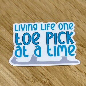 May include: White sticker with a blue and gray ice skate design. The sticker says "Living life one toe pick at a time."