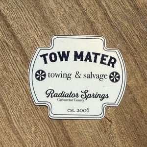 May include: A white and black sticker with the text "TOW MATER" above "towing & salvage" and "Radiator Springs" with "Carburetor County" and "est. 2006". The sticker has a unique shape and is on a wooden surface.