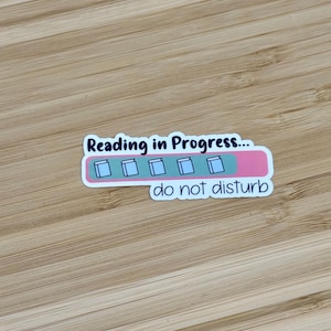 May include: A sticker with a pink and white progress bar that reads "Reading in Progress... do not disturb". The progress bar is filled with five white squares, each representing a book.