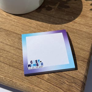 May include: A square notepad with a gradient of blue and purple. The notepad has the text "CAST MEMBER YOU ARE THE MAGIC" in blue and white. The notepad is on a wooden surface.