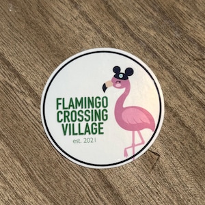 May include: A round white sticker with a black border features a pink flamingo wearing a black hat. The text "FLAMINGO CROSSING VILLAGE" is in green, with "est. 2021" below. The sticker is on a wooden surface.