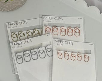 May include: Four sets of five paper clips in different shapes and colors. The first set is gold star-shaped paper clips. The second set is rose gold heart-shaped paper clips. The third set is silver heart-shaped paper clips. The fourth set is silver star-shaped paper clips. Each set is packaged in a clear plastic sleeve with a white card that says "PAPER CLIPS" and "5-pieces".