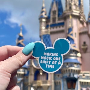 May include: A blue and white sticker with Mickey Mouse ears that says "Making magic one shift at a time". The sticker is held in front of a blue and pink castle.
