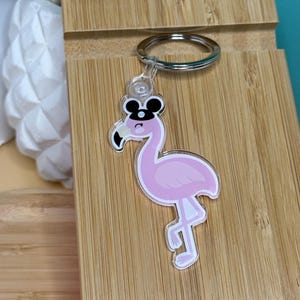 May include: Pink flamingo keychain with black Mickey Mouse ears and an eye patch. The keychain is made of clear acrylic and has a silver metal ring.