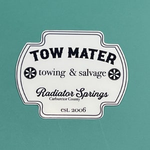 May include: A white sticker with a black border and text that reads "TOW MATER" above "towing & salvage" and "Radiator Springs" with "Carburetor County" and "est. 2006". The sticker has a teal background.