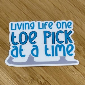 May include: A white sticker with the text "Living Life one toe PICK at a time" in blue lettering. The sticker has a light gray figure skating blade design at the bottom, and is placed on a wooden surface.