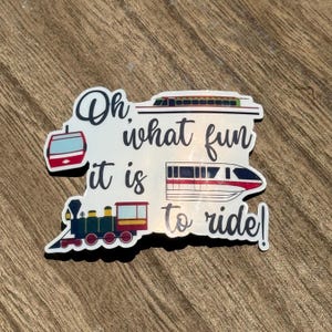 May include: A white sticker with the text "Oh, what fun it is to ride!" and illustrations of a gondola, a monorail, and a train. The sticker has a black border and is on a wooden surface.