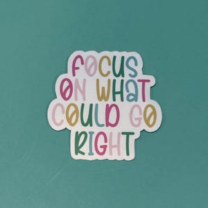 May include: A white sticker with the phrase "FOCUS ON WHAT COULD GO RIGHT" in colorful, bubble-style letters. The sticker has a rounded, cloud-like shape and is set against a teal background.