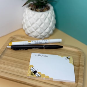 May include: A white notepad with a bee and honeycomb design. The notepad has the text "Bee positive" written on it. The notepad is on a wooden tray with a yellow pineapple plant in the background.