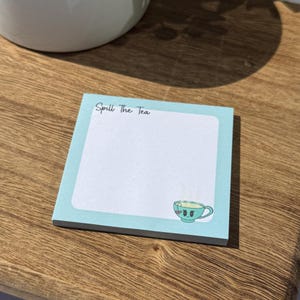May include: A square notepad with a light blue border and the text "Spill The Tea" at the top. A cartoon teacup with a smiling face is in the bottom right corner. The notepad is on a wooden surface.