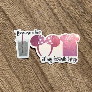 May include: A sticker featuring a drink, Minnie Mouse ears, and a backpack. The text reads "These are a few of my favorite things." The drink is in a clear cup with a straw. The ears and backpack are pink with white polka dots.