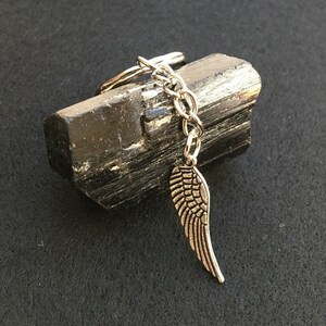 May include: A silver-coloured keyring featuring a wing charm suspended from a chain. The wing charm is detailed with feather-like lines. The keyring rests on a dark, textured stone, against a black background.