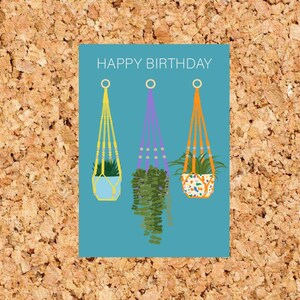 May include: A blue card with the text "HAPPY BIRTHDAY" and three hanging plants with colourful pots. The plants are a green succulent, a purple plant, and a green plant with a patterned pot.