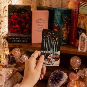 Puede incluir: Una carta de tarot Diez de Espadas sostenida verticalmente, con una ilustraci&oacute;n de diez espadas atravesando una figura. La carta se muestra entre varios mazos de or&aacute;culo, incluyendo Message of Love, Soulmate Love Message, The Light Seer's y Wisdom of Oracle.