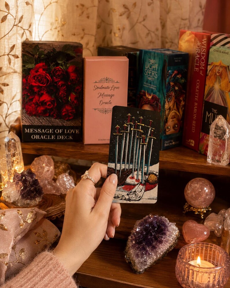 Puede incluir: Una carta de tarot Diez de Espadas sostenida verticalmente, con una ilustraci&oacute;n de diez espadas atravesando una figura. La carta se muestra entre varios mazos de or&aacute;culo, incluyendo Message of Love, Soulmate Love Message, The Light Seer's y Wisdom of Oracle.