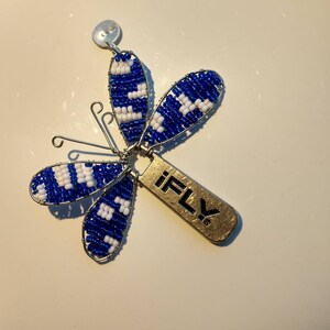 Könnte beinhalten: Ein blauer und weißer Perlen-Schmetterlings-Anhänger mit einem silbernen Drahtrahmen und einem goldenen Anhänger mit der Aufschrift "iFLY".