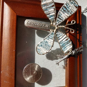 Könnte beinhalten: Ein silberner und blauer Perlen-Schmetterlingsanhänger mit dem Wort "DESTINATION" auf einem silbernen Anhänger, der an einer silbernen Kette mit einem kleinen Flugzeug-Anhänger hängt. Der Anhänger wird in einem Holzrahmen mit einem US-Penny ausgestellt.