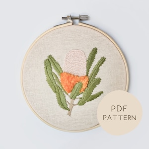 May include: Embroidered hoop art featuring a pink and orange flower with green leaves. The hoop is made of wood and has a beige linen fabric. The text "PDF PATTERN" is printed on a beige circle.
