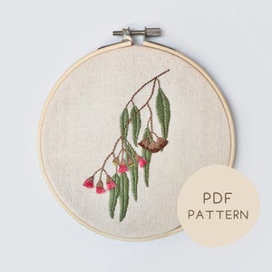 May include: Embroidered eucalyptus branch with green leaves and pink flowers, displayed in a wooden embroidery hoop. The design is stitched on a beige fabric. A circular label with the text "PDF PATTERN" is visible.